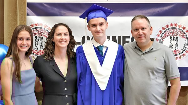 Un grupo de cuatro personas posan en la ceremonia de graduación en el campus de la Creston Valley Secondary School con un joven en toga y birrete.