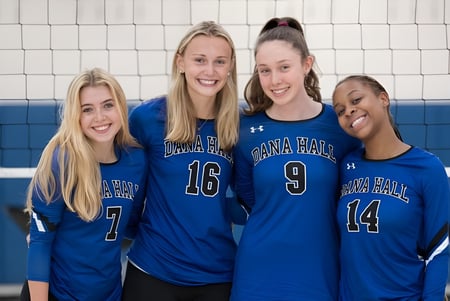 Cuatro estudiantes de la Dana Hall School están en uniformes de voleibol en el gimnasio frente a una red de voleibol.