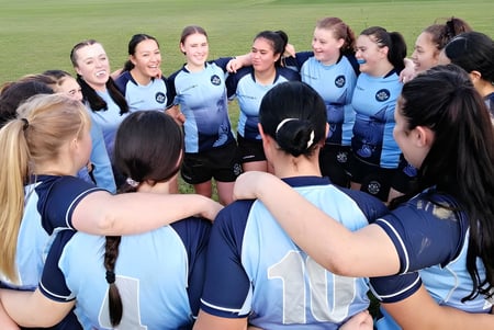 Un grupo de atletas femeninas de la Dannevirke High School en uniformes azules y blancos se consulta en el campo de deportes.