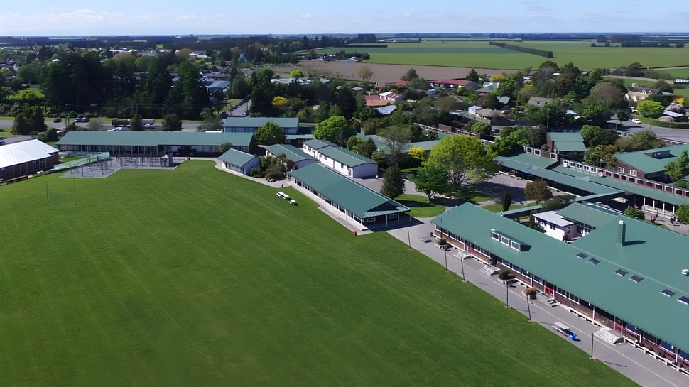 Paisaje con campos verdes y edificios frente a colinas boscosas en el terreno de la Darfield High School.