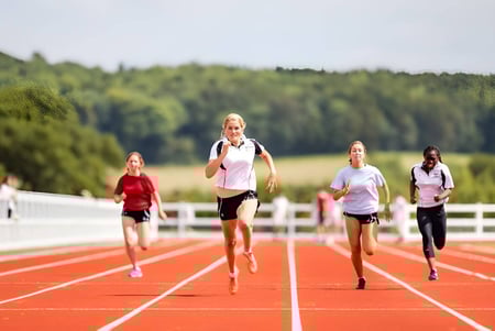 Un grupo de corredoras sprinta en la pista en el campo deportivo de Dauntsey’s School frente a un fondo boscoso.