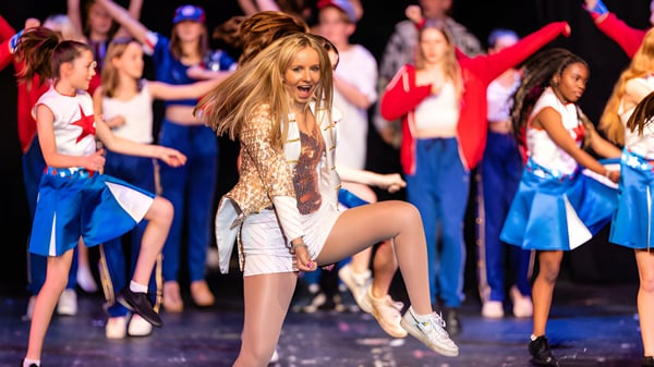 Una estudiante de la Dauntsey’s School baila con un atuendo brillante en el escenario con un grupo de bailarinas y bailarines en trajes azules y rojos.