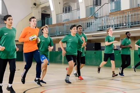 Un grupo de estudiantes del David Game College Liverpool corre juntos en el gimnasio con cancha de baloncesto y gradas.