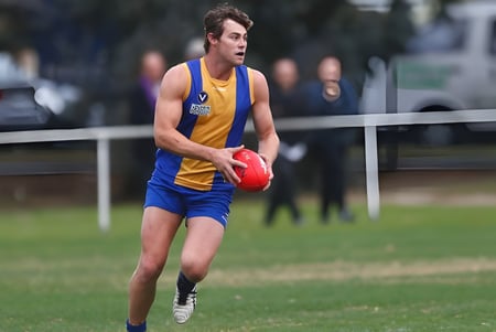 Un joven atleta en uniforme azul y amarillo corre con un balón de fútbol rojo en el campo del De la Salle College Churchtown.