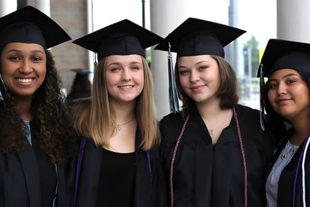 Cuatro graduadas en toga y birrete celebran su graduación frente a una ventana en el campus de Deer Valley Unified.