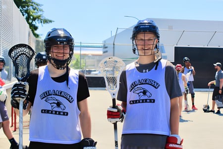 Dos alumnas de la Delta Secondary School llevan uniformes de lacrosse y están en un campo deportivo.