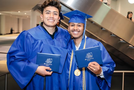 Dos estudiantes del Denver Public School District están en el pasillo con toga de graduación azul y sosteniendo orgullosamente sus diplomas.