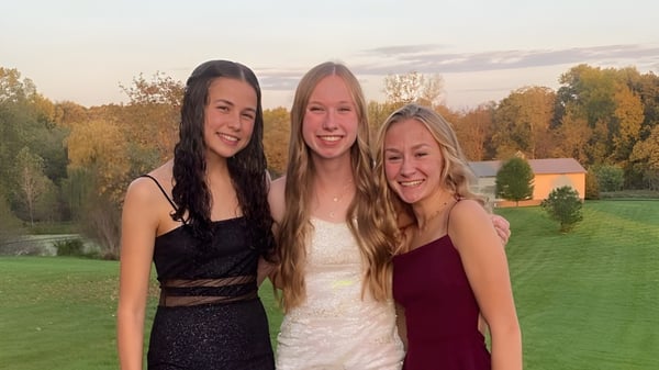 Tres estudiantes de la Dexter High School posan juntas con ropa formal frente a un prado verde con árboles.