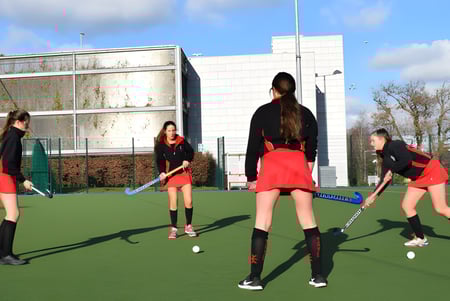 Las alumnas del Dominican College Sion Hill juegan al hockey sobre césped en el campo de césped artificial con un edificio escolar al fondo.