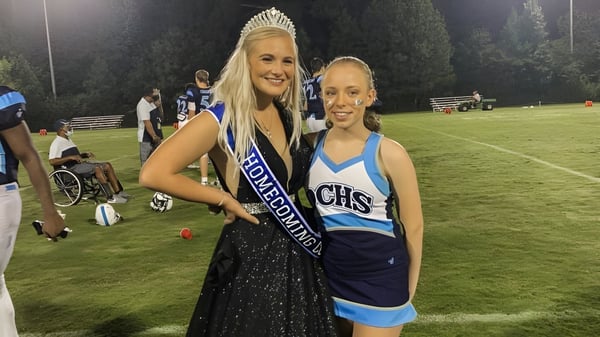 Dos estudiantes de la Dominion Christian School están de pie por la noche en el campo deportivo, una como Reina de Homecoming con corona y banda, la otra en uniforme de animadora.