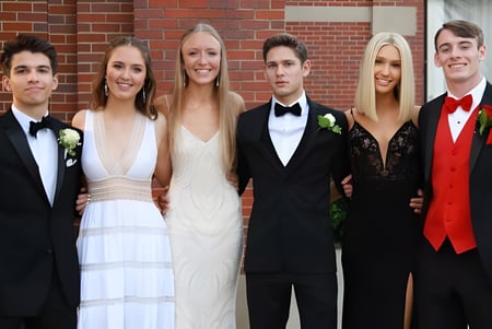 Un grupo de personas bien vestidas, entre ellas una pareja en ropa formal, posan frente a un edificio de ladrillo en el campus del Don College.