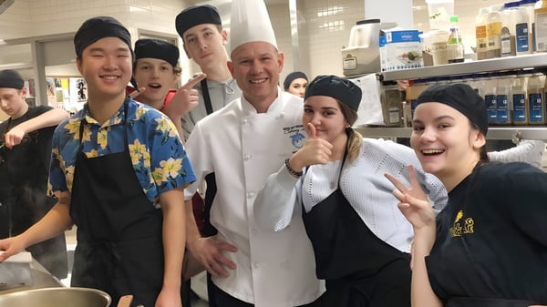 Alumnos de Dover Bay Secondary School se reúnen en la cocina de la escuela con un chef en uniforme blanco.