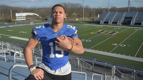 Una persona con camiseta deportiva azul está en el campo de fútbol del Downingtown Area School District con el estadio y las gradas al fondo.