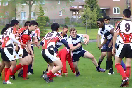 Estudiantes del Dr. Norman Bethune Collegiate Institute juegan al rugby en un campo deportivo con casas residenciales de fondo.