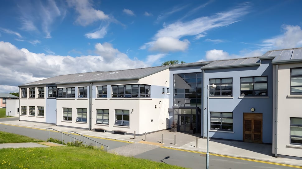 El moderno edificio de varios pisos de la Drogheda Grammar School con grandes ventanas y un campo en primer plano bajo un cielo azul.
