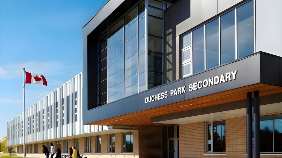El moderno edificio de varios pisos de la Duchess Park Secondary School con grandes ventanas de vidrio bajo un cielo azul y una bandera canadiense en primer plano.