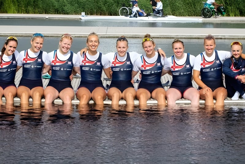 Un grupo de atletas de la Durham School está sentado en el agua con hierba y ciclistas al fondo.