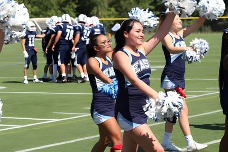 Las animadoras de la East Catholic High School realizan una actuación en el campo de fútbol con jugadores al fondo.