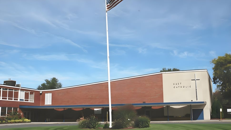 El edificio de ladrillo de la East Catholic High School con una gran bandera americana frente a un cielo azul.