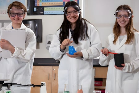Tres alumnas con batas de laboratorio blancas están en el laboratorio de la École Dansereau Meadows School con equipo científico.