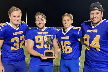Cuatro estudiantes de la École Sainte-Anne llevan camisetas de fútbol y sostienen un trofeo en el campo de fútbol de noche.