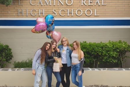 Cuatro estudiantes están frente al edificio de la El Camino Real Charter High School sosteniendo globos de colores.
