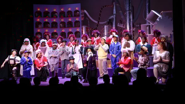 Un gran grupo de estudiantes de Elgin Park Secondary School está de pie en coloridos disfraces en el escenario con un elaborado decorado y iluminación.