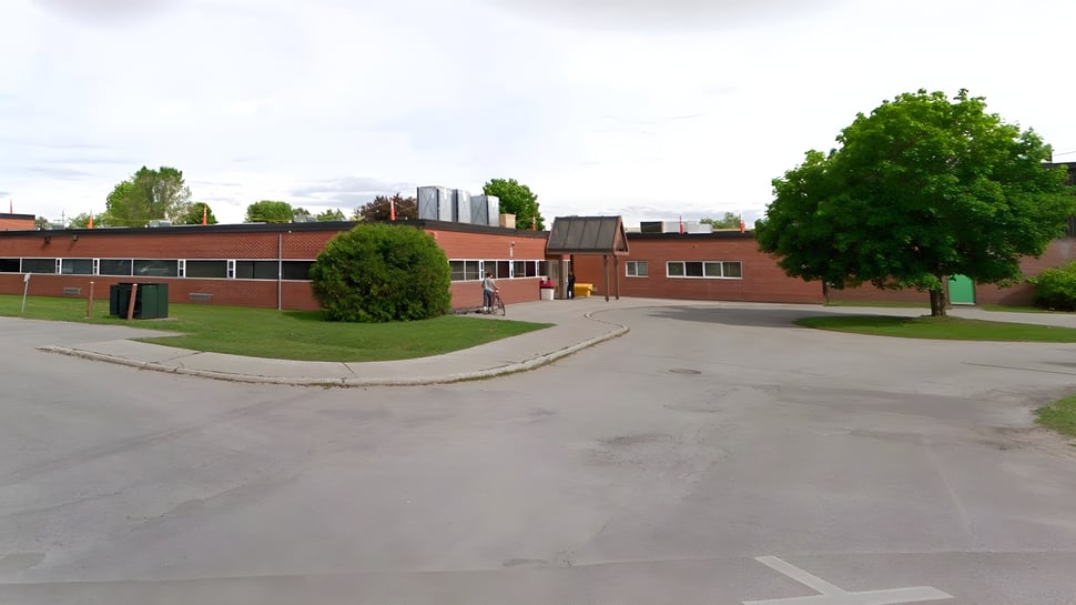 El edificio de ladrillo de la Elmvale District High School con pradera y árboles en primer plano está rodeado por un camino pavimentado y otros edificios al fondo.
