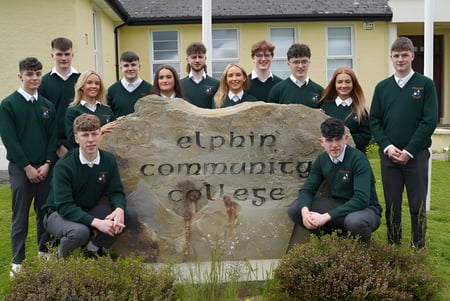 Un grupo de estudiantes en uniformes escolares posan frente al gran letrero de piedra del Elphin Community College.