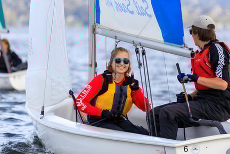 Dos personas están sentadas en un pequeño velero sobre el agua con una vela azul y blanca de fondo en el terreno de la Embley.