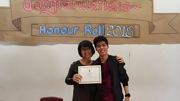 Dos personas sostienen juntas un certificado frente a un banner en el campus del Emery Collegiate Institute.