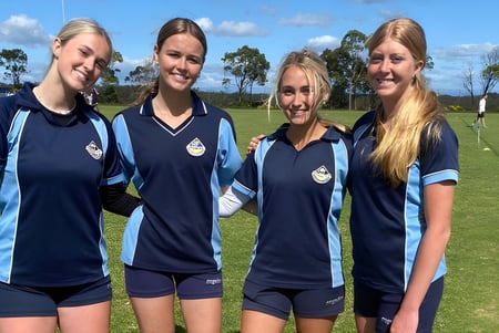 Cuatro estudiantes de la Engadine High School están juntas en un campo deportivo con árboles y un cielo azul de fondo.
