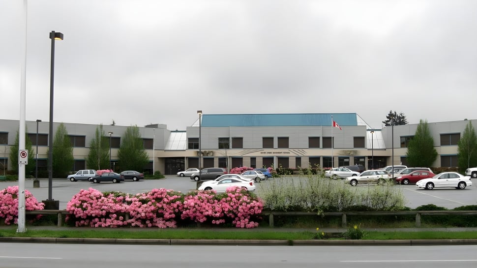 El edificio de la Enver Creek Secondary School con un estacionamiento en el frente y flores rosas floreciendo bajo un cielo nublado.