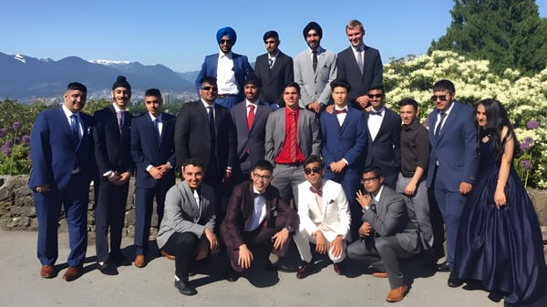 Un grupo de hombres en ropa formal posan afuera con un paisaje montañoso de fondo en el terreno de la Enver Creek Secondary School.