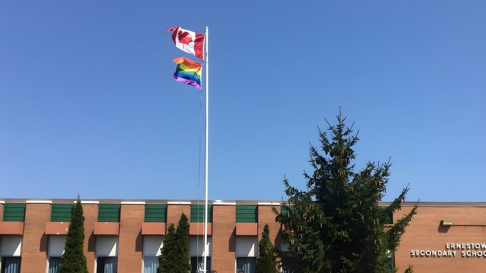 Frente al edificio de ladrillo de la Ernestown Secondary School ondean una bandera canadiense y una bandera del arcoíris en el mástil.