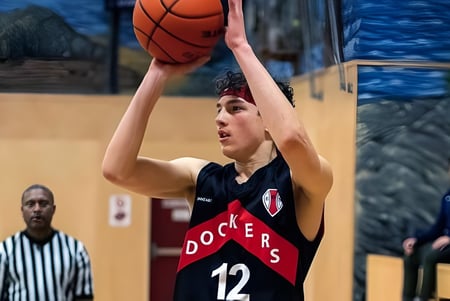 Un estudiante de la Esquimalt High School salta para lanzar a canasta en la cancha de baloncesto con espectadores al fondo.