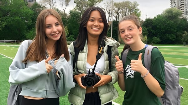 Tres estudiantes están sonriendo en el verde del campus del Etobicoke Collegiate Institute y cada una muestra un pulgar hacia arriba.