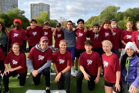 Un grupo de estudiantes del Etobicoke Collegiate Institute lleva camisetas burdeos y está junto en el parque frente a edificios altos.