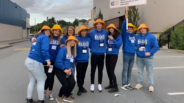 Un grupo de estudiantes de la Exploits Valley High School está frente a una obra de construcción con camisas azules y cascos de seguridad.
