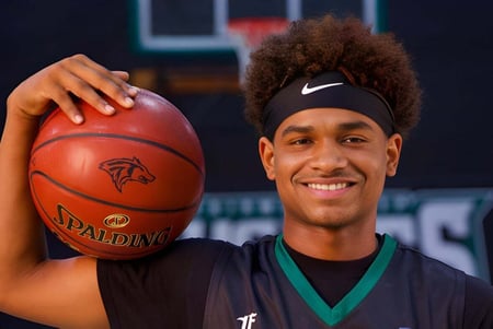 Un estudiante de la Fairmont Prep Academy con rizos y una banda de Nike sostiene un baloncesto frente al campo de baloncesto.