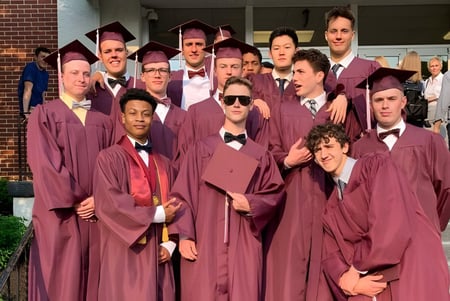 Un grupo de graduados de la Faith Christian Academy (PA) está de pie en una toga roja frente a un edificio de ladrillo.