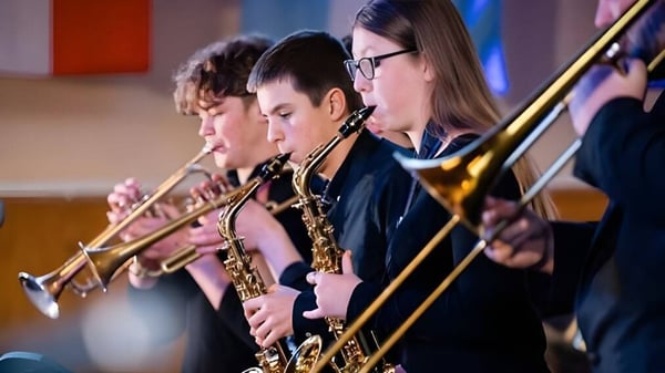 Un grupo de músicos toca en el escenario de la Felsted School diversos instrumentos de metal y de viento frente a un fondo colorido.