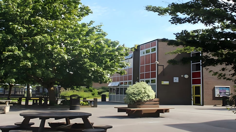 El patio de la Ferndown Upper School muestra bancos de madera frente a un edificio de ladrillo y está rodeado de árboles.