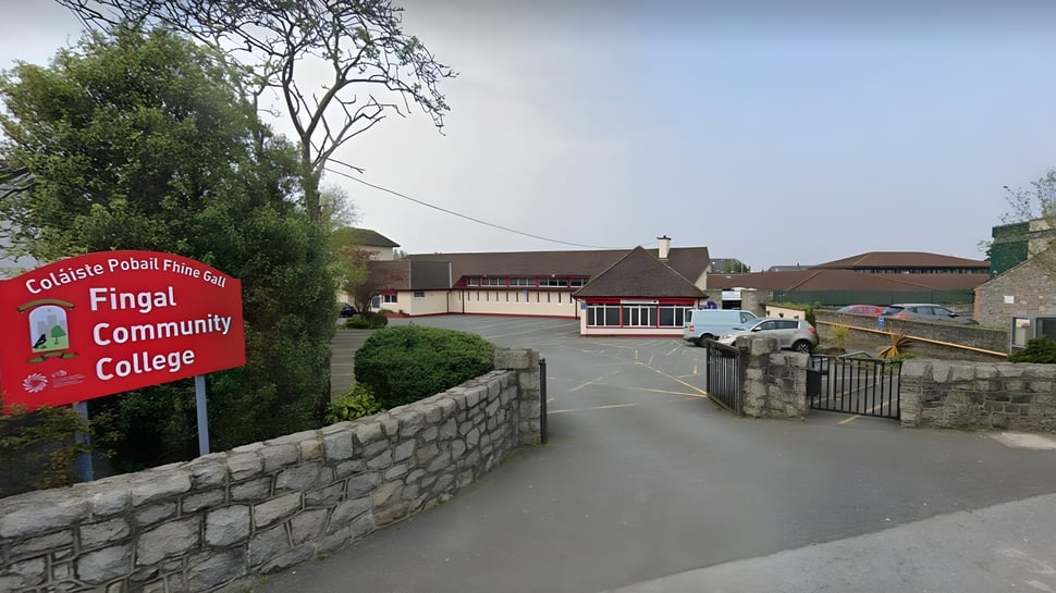 Un cartel rojo del Fingal Community College está frente a una pared de piedra con edificios al fondo.