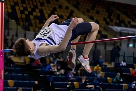 Un atleta masculino de la Firrhill High School salta en el salto de altura sobre una barra en un gimnasio con espectadores.