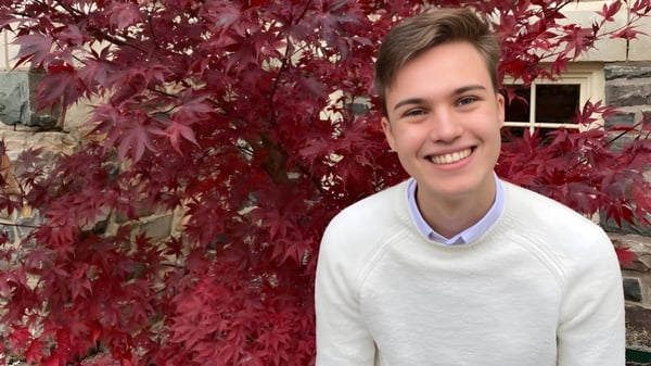 Una persona con un suéter blanco está de pie frente a las hojas otoñales en el terreno de la FJ Brennan Catholic High School.