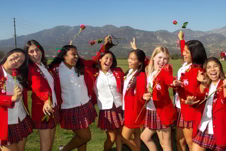 Estudiantes de la Flintridge Sacred Heart Academy celebran en togas de graduación rojas y blancas frente a un paisaje montañoso.