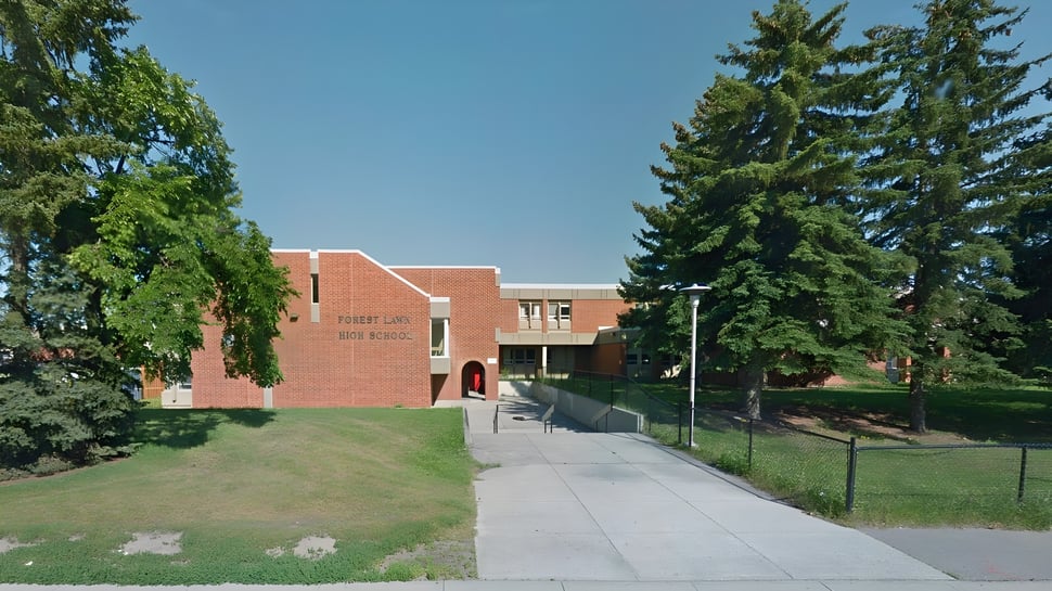 El edificio de ladrillo de la Forest Lawn High School con techo rojo está rodeado de árboles verdes bajo un cielo azul.
