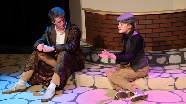 Dos personas con disfraces coloridos están sentadas en el escenario de la Forsyth Country Day School con una pared de ladrillo de fondo.