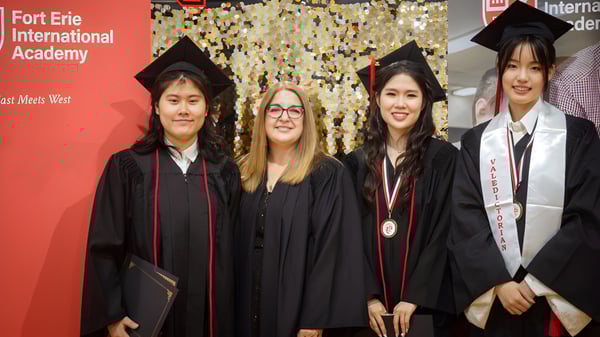 Un grupo de graduadas y graduados de la Fort Erie International Academy posan con vestimenta académica frente a un banner con el nombre de la escuela.